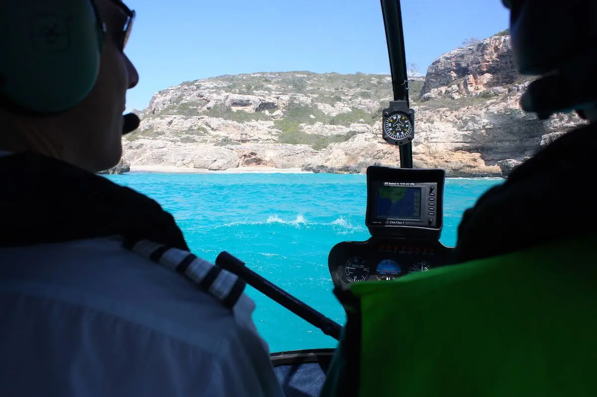 Pilots flying a helicopter towards sea cliffs over turquoise water
